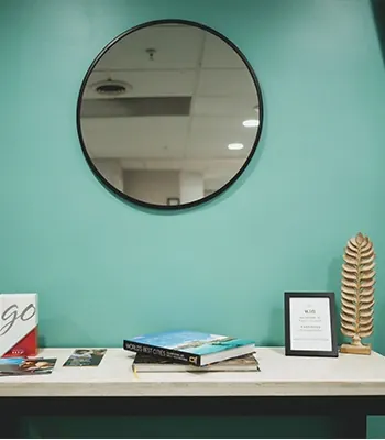 A round mirror on the teal wall above a console table with magazines.